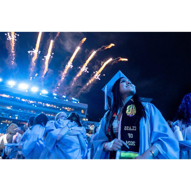 Graduate in cap and gown looks up at fireworks over Kenan Stadium during Spring Commencement 2024.